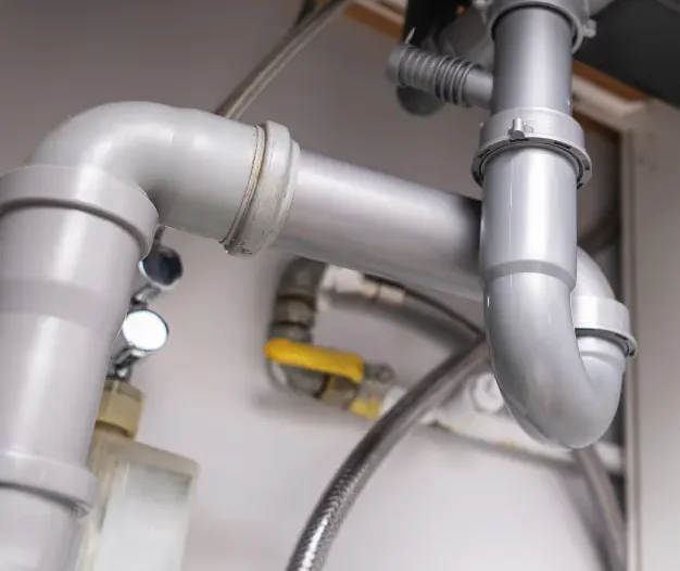 A close up view of sink plumbing showing white PVC pipes, a black garbage disposal unit, and various water lines and connections under a